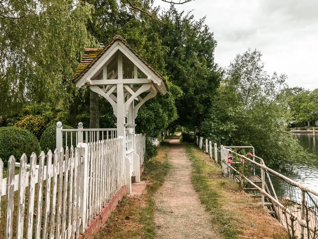 A thin trail leading ahead, lined with a white fence on the left, and metal railings and river to the right. The path is lined with trees ahead.