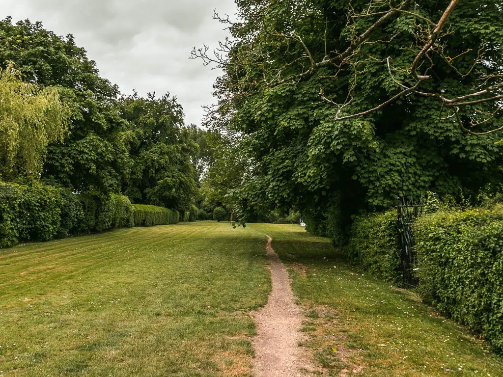 A thin trail leading through a neatly cute grass lawn. There are hedges lining the edges of the lawn, with big trees behind the hedges.