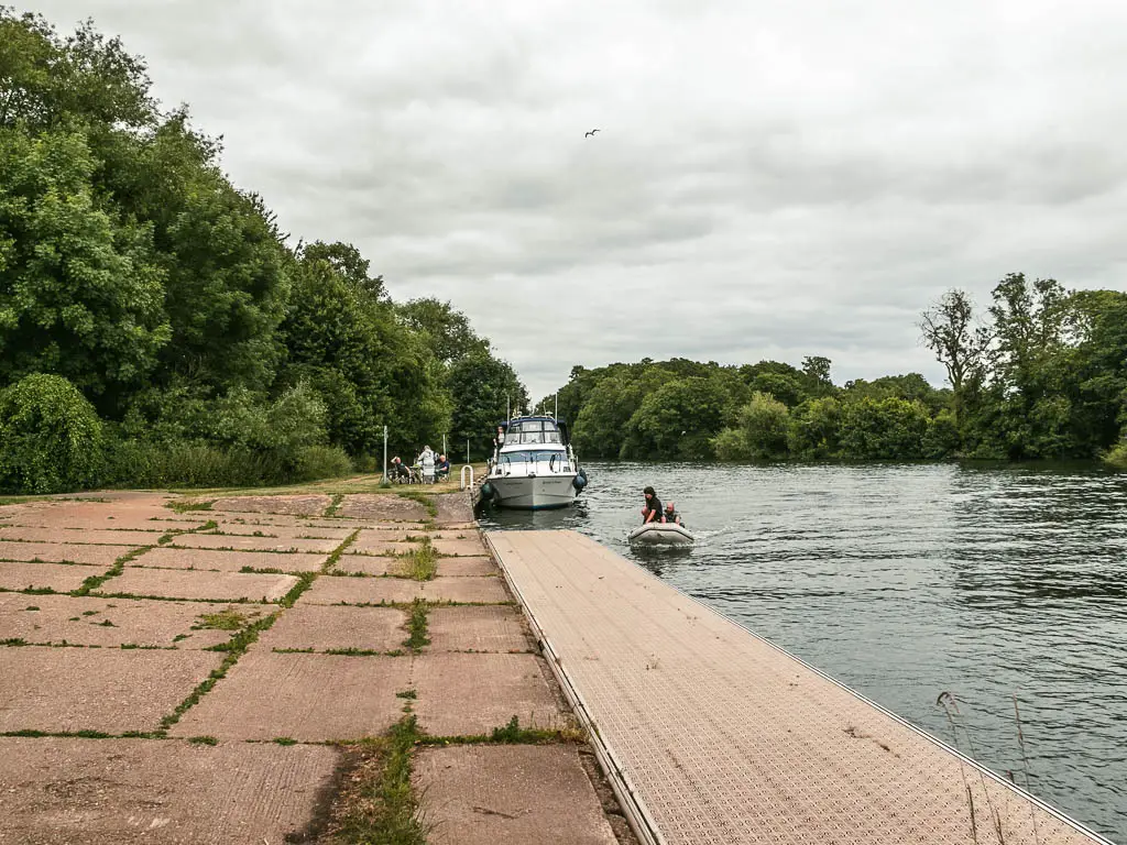 A large open concrete area, with the river to the right. There is a big boat moored to the side, and a small dingy boat with people in it.