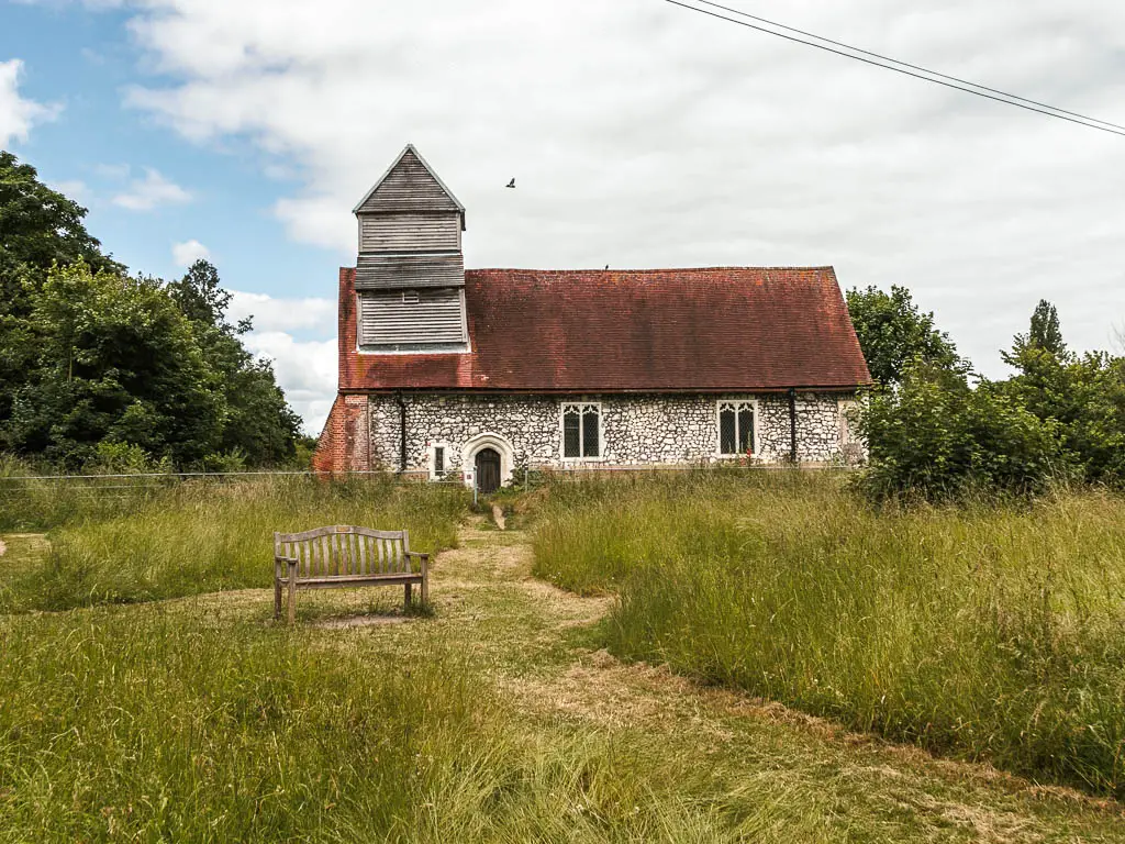 A stone walked church with ed roof on the other side of a meadow with tall green grass and a strip of cut grass forming a path. There is a wooden bench next to the path.