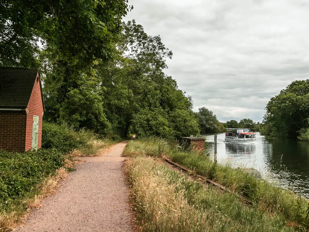 A gravel path leading ahead on the left, with a strip of unkempt grass on the right then the river, on the walk from Maidenhead to Windsor. There is a big boat on the river. There are trees lining the path ahead.