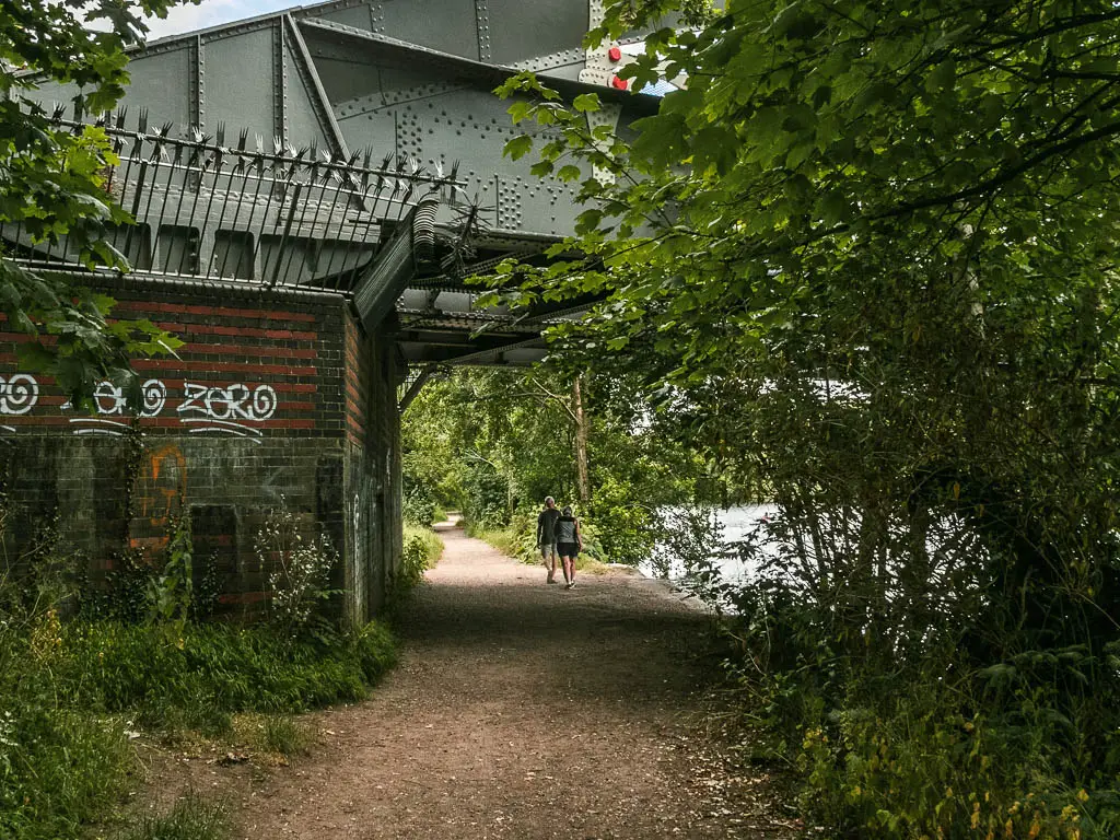 A path leading under a metal bridge with a graffiti covered brick wall to the left. There are two people walking under the bridge.