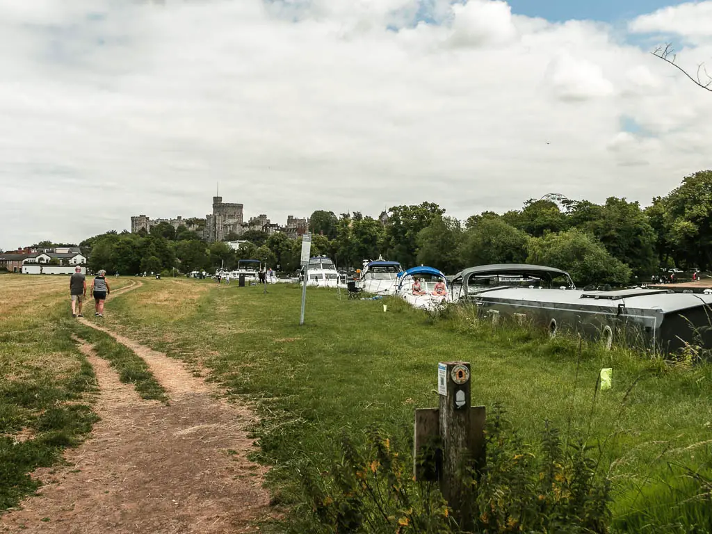 A dirt trail leading through a large open grass field with a view to Windsor castle behind the tree tops ahead in the distance, at the end of the walk from Maidenhead. There are boats lining the side of the river all the way along. There are two people walking on the trail ahead.