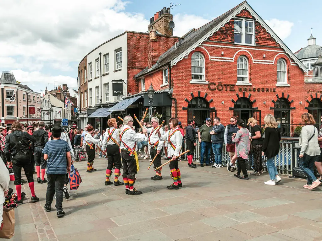 Lots of people and morris dancers standing on the paved ground with shop buildings ahead, at the end of the walk in Windsor.