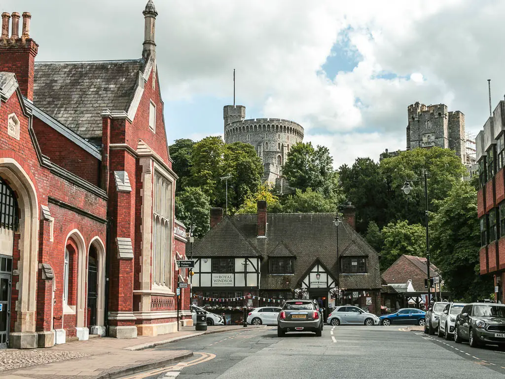 Looking along the road, with a view of the top of Windsor castle behind the trees ahead. There is a Tudor building  below the trees, and a red bricked building on the side of the road to the left. There is a car driving along the road.