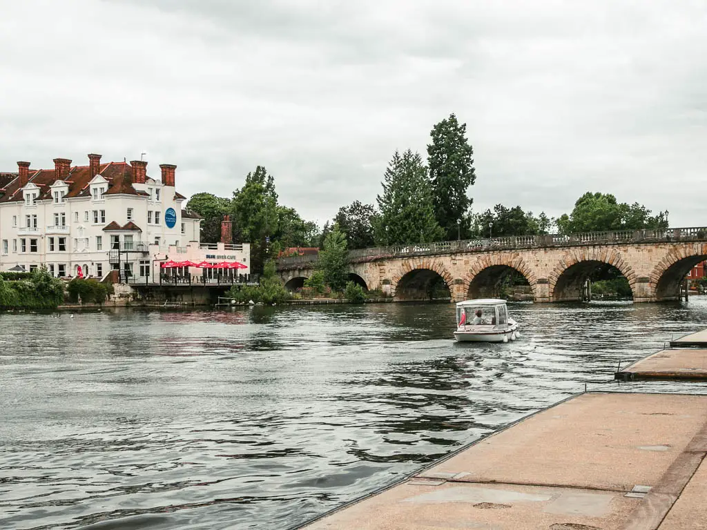 The river with ripples from the small boat, heading towards an archway bridge. There is a walkway on the right. There is a big white walled building on the other side of the river.