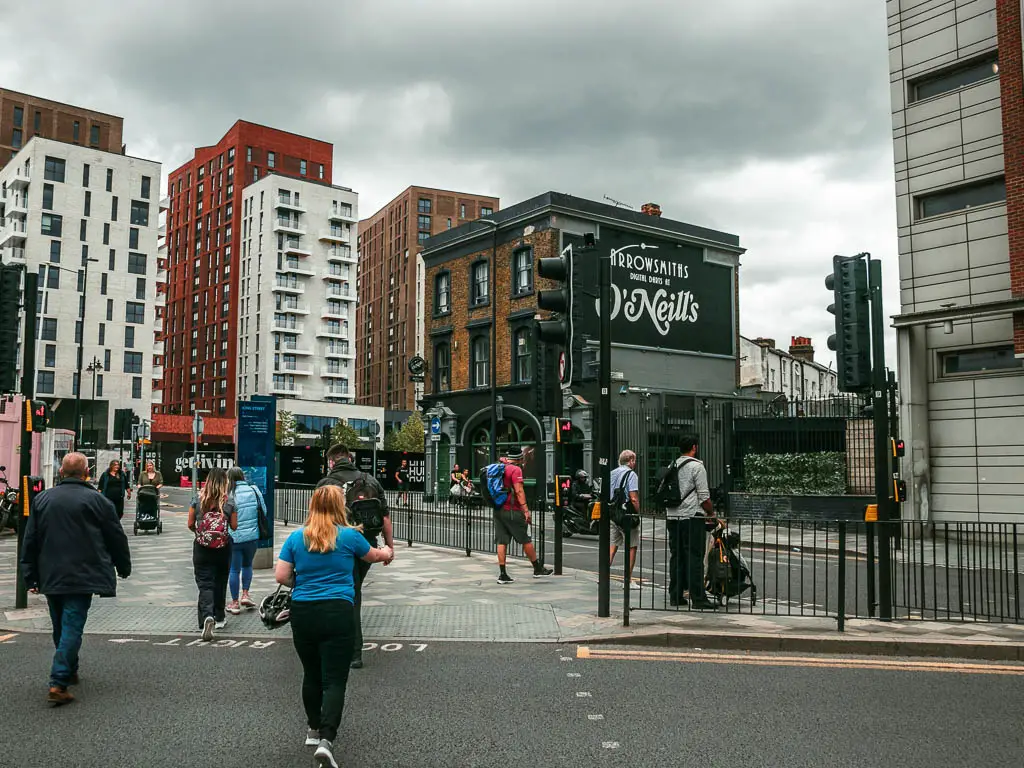 Walking across the road to the central reservation, with masses of other people. There are tall apartment buildings on the other side. It's a grey day. 