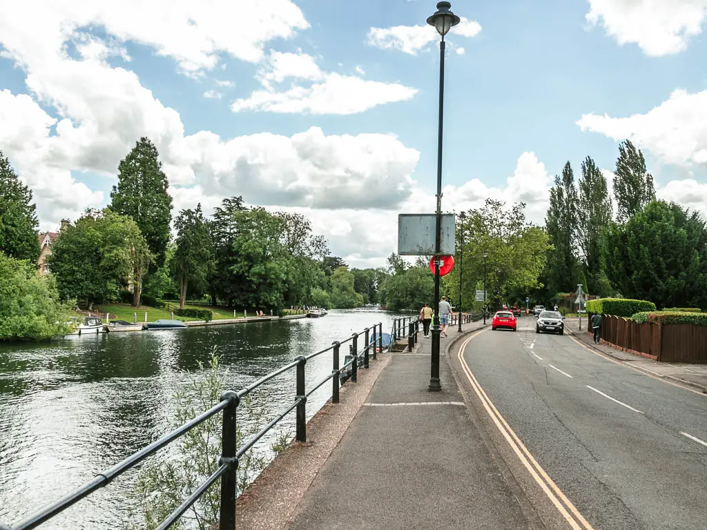 A pavement leading ahead, with the road to the right and metal railing and river to the left. 