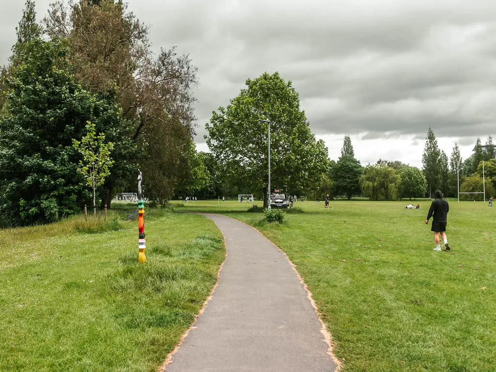 A path leading through a large sports field. There is a mad wearing black jumper walking in the field. There are trees lining the path ahead.