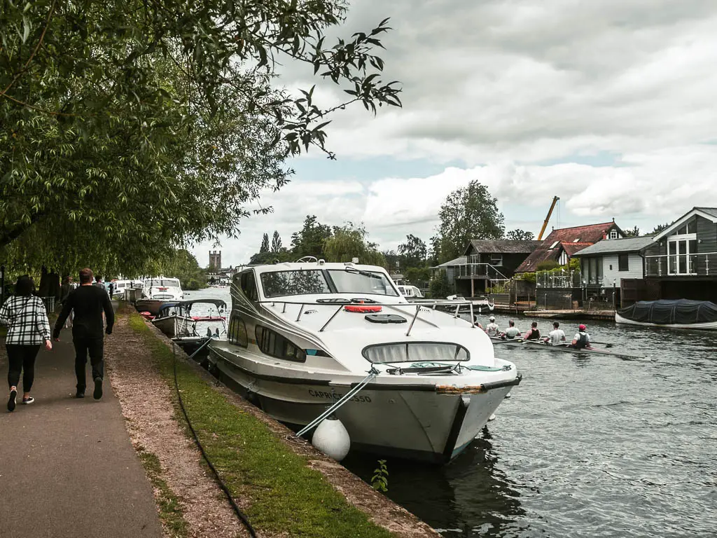 A path on the left, with the river on the right. There are speed boats moored to the side, rowers in the water, and people walking along the path.