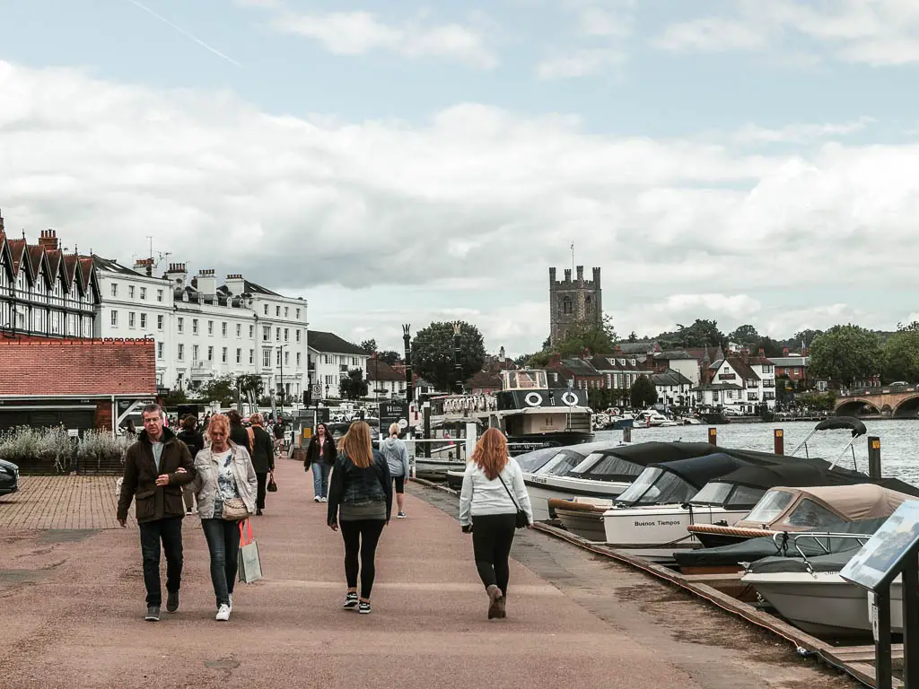 A mass of people walking along the wide path, with the river on the right and lots of boats moored to the side. There are the houses and buildings of Henley ahead.