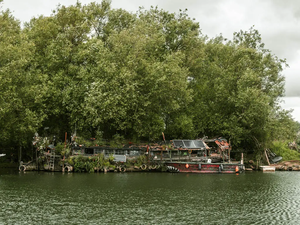 Looking across the river with ripples, to a scruffy house boat on the other side, at the start of the walk from Reading to Henley. There is a mass of green leafy trees behind the boat and hanging over it.