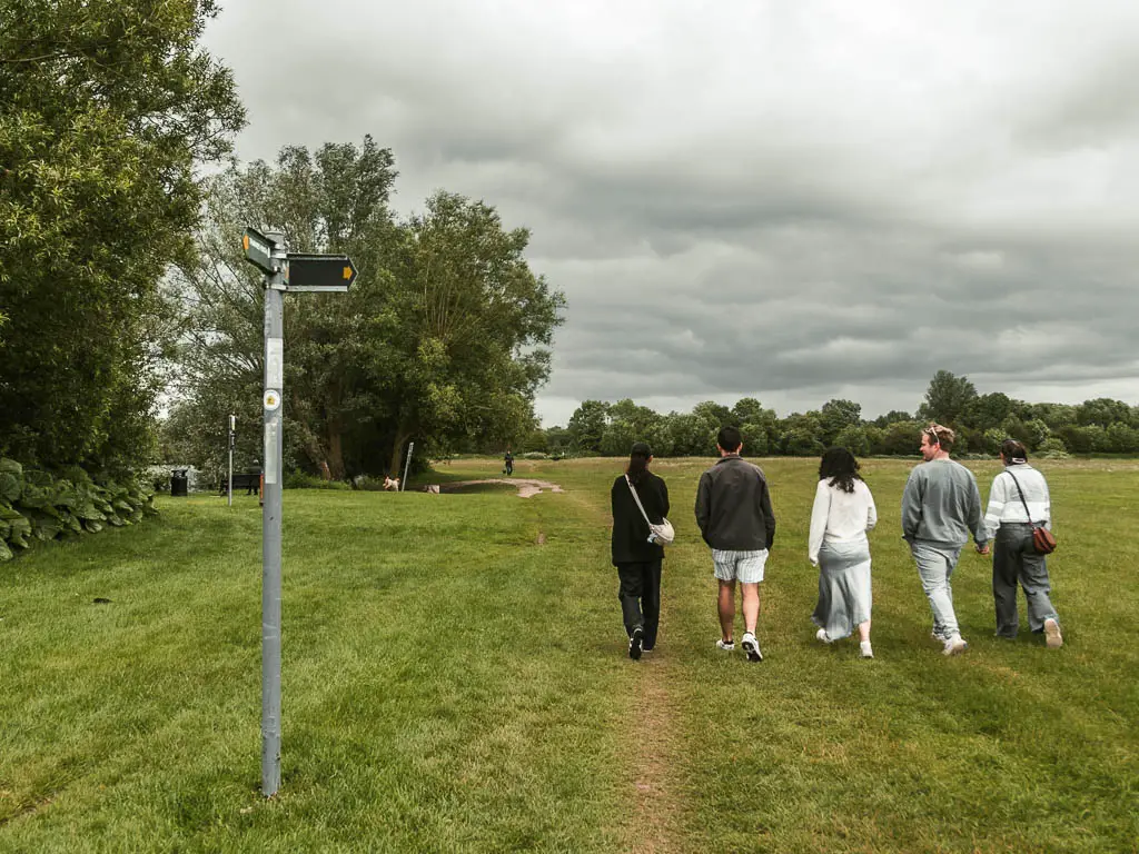 Five people in a row walking across a large grass field. There is a trail signpost on the left.