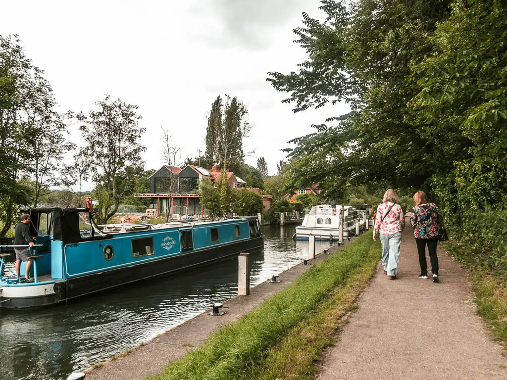 A path on the right and river on the left, with a blue barge heading towards the lock. There are two women walking on the path.