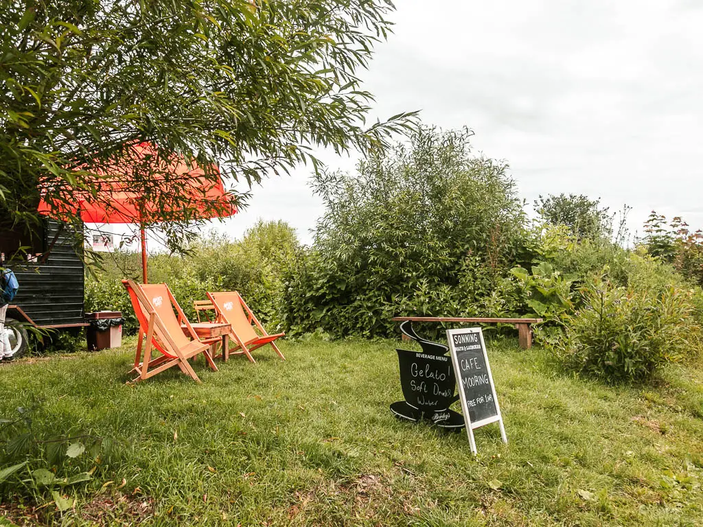 Two orange deck chairs and umbrella on a small green grass hill, and a black information board in front of them.