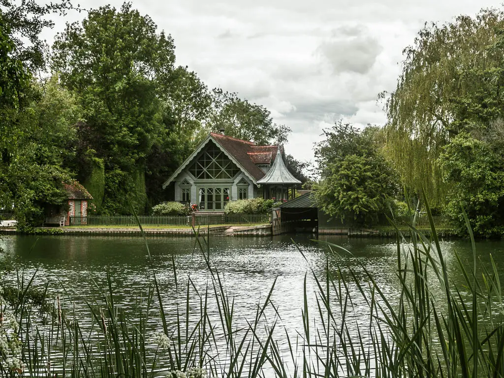 Looking over the tab, grass to the river and small house on the other side, on the walk from Reading to Henley. The house is surrounded by lots of trees of varying sizes. 