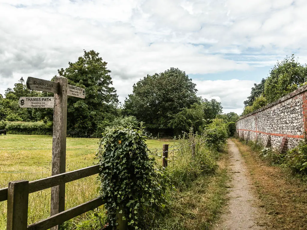 A path leading ahead, with a stone wall on the right and wooden fence on the left. There is a wooden trail signpost next to the fence and a grass field on the other side.