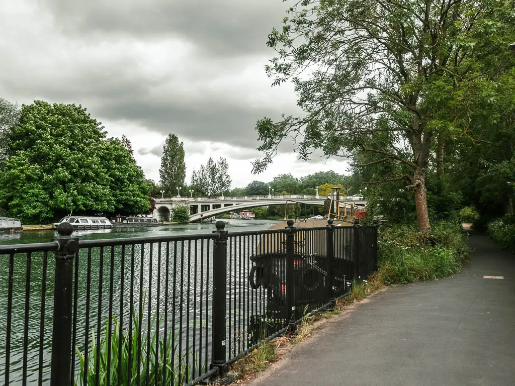 The path on the right, with black metal railings on the left and the river on the other side. There is a white bridge in the distance over the river, leading into a big bushy tree on the other side.