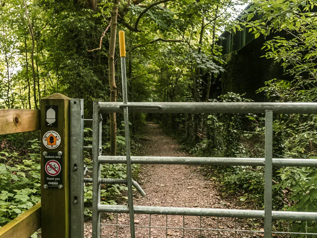 a metal gate with a white acorn and yellow arrow on the left wooden pole. The arrow points ahead to the dirt path. The dirt path is lined with bushes and trees.