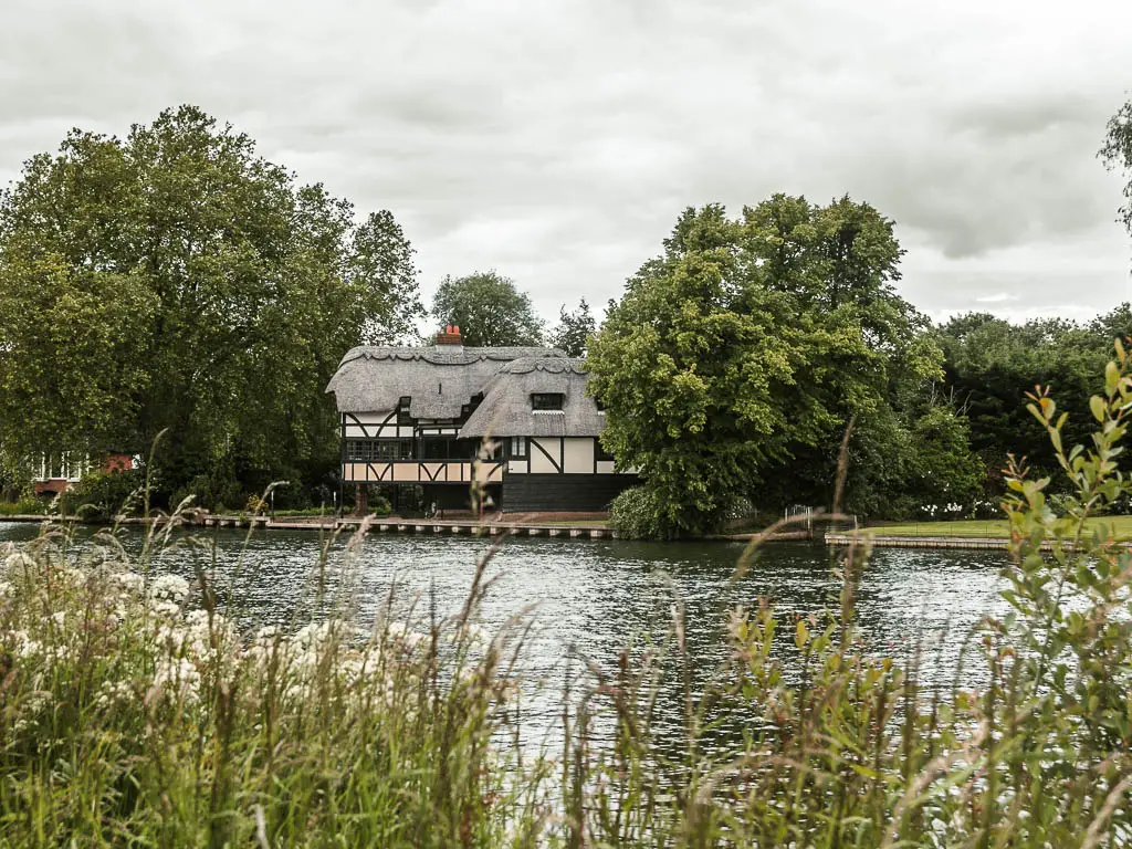 Looking over the tall grass to the river with ripples and Tudor style building on the other side. The building is surrounded by big trees with masses of green leaves.