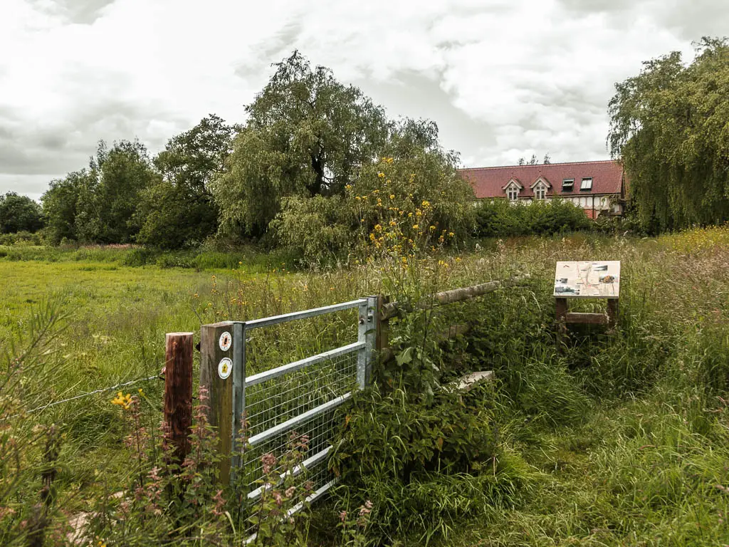 A metal gate on the left with a field on the other side. The gate and fence is partially hidden by bushes. There is the rooftop of a hose visible ahead behind the bushes and trees.