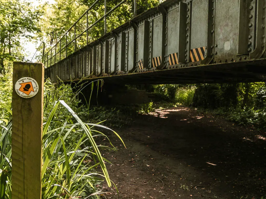 A wooden stump signpost on the left, with a yellow arrow pointingt ahead to the right under a low metal bridge.