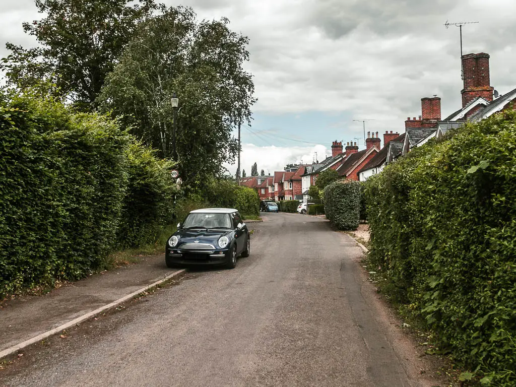 A road leading ahead, lined with hedges on both sides. There is a pavement on the left with a car parked half on it. There is a row of houses along the right side of the road.