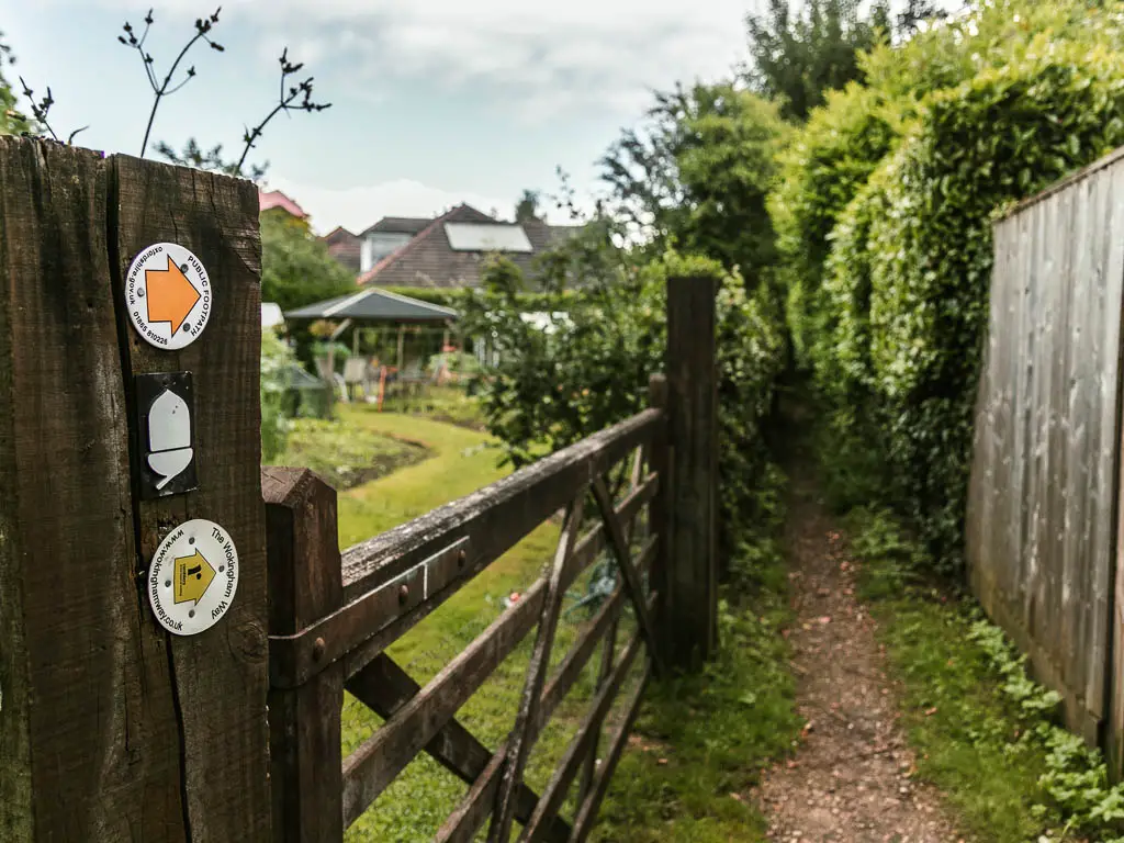 A narrow dirt trail with a wooden fence and garden to the left and wooden fence then hedge to the right. There is a white acorn and arrange arrow pointing along the trail, on the wooden fence on the left.