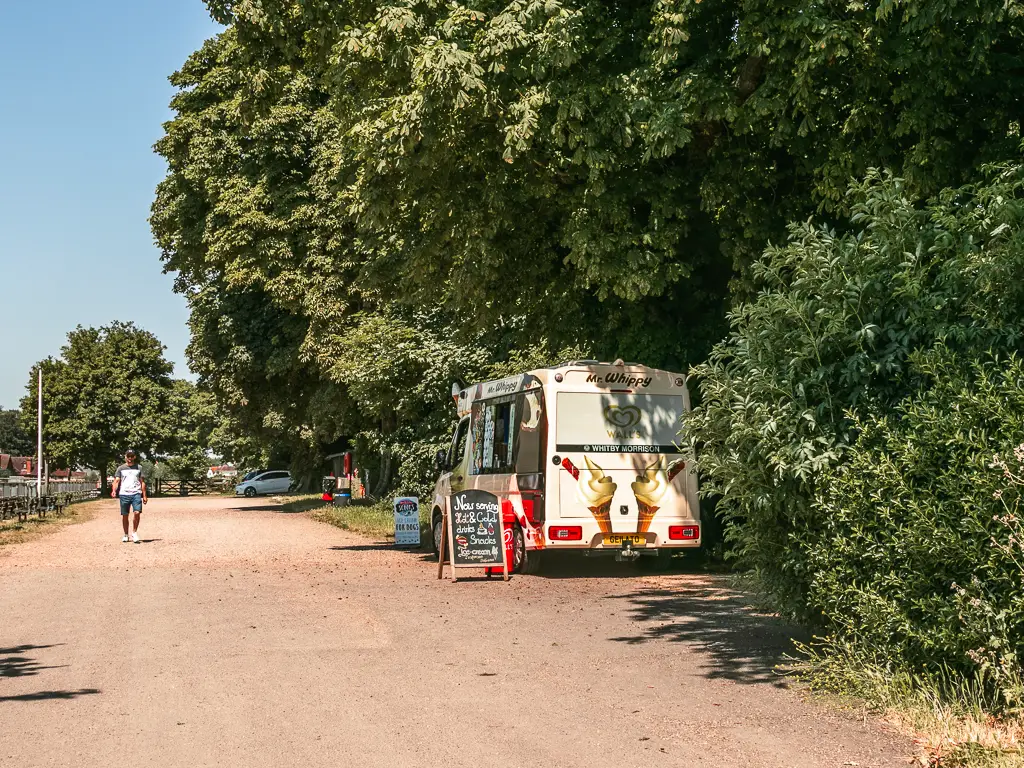 An ice cream van nestled under the trees on the side of the wide road. 