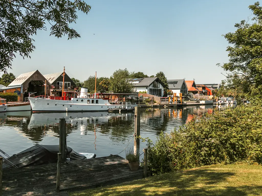 Looking over the neatly cut grass to the river with houses on the other side, on the walk between Staines and Shepperton. There is a big white boat moored to the other side.