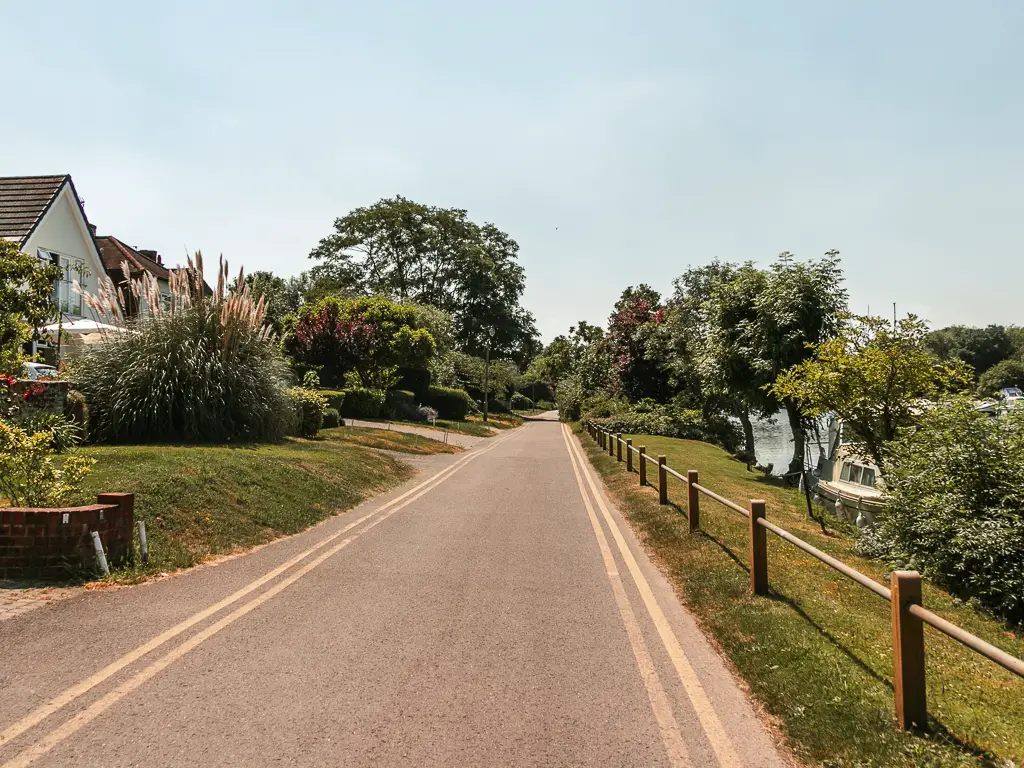 A road leading ahead, lined with strips of grass, with houses and bushes on the left, and the river just visible on the right.