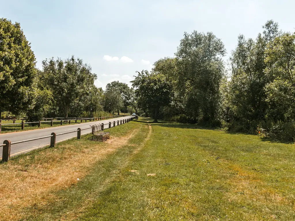 A large green with railing and road on the left, and big trees ahead. There is a metal and wooden bench on the green.