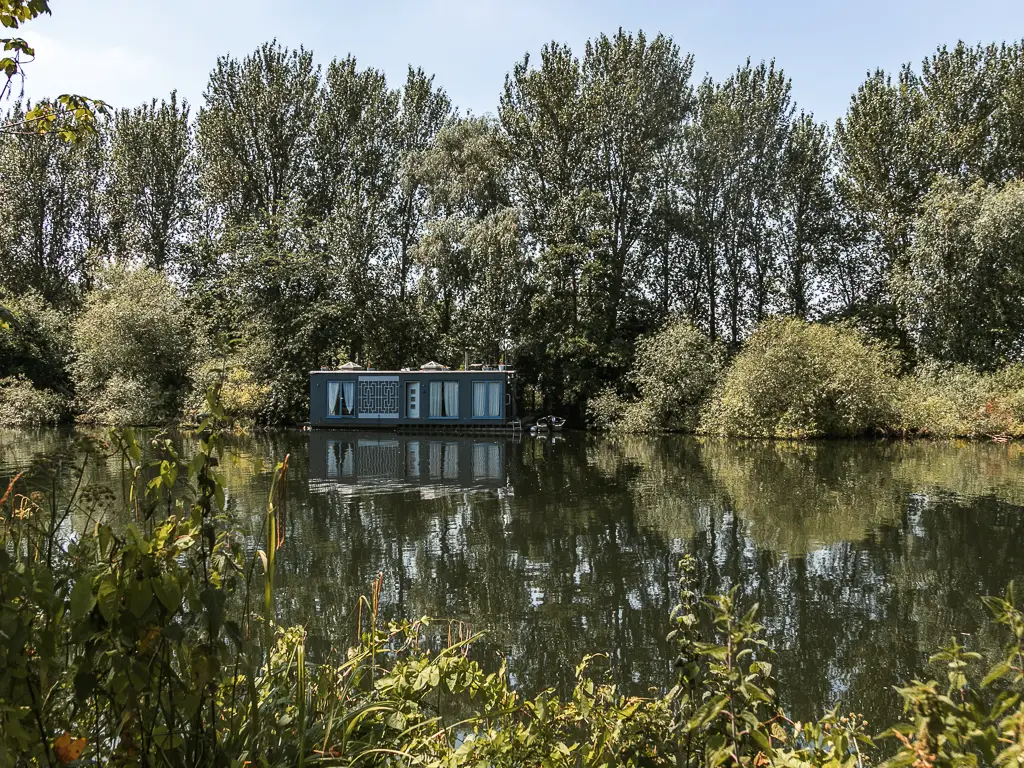 Looking over the bushes to the river and small house boat on the other side with bushes and tall trees behind it, on the walk from Staines to Shepperton.