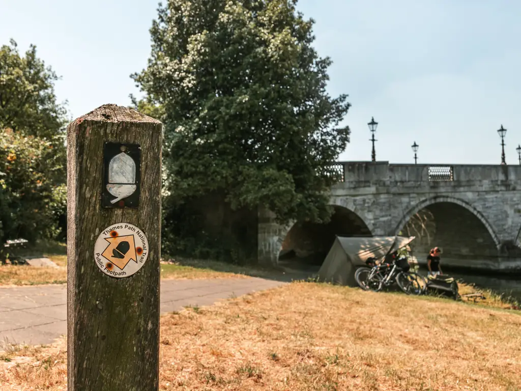 A wooden trail sign post with white acorn and yellow arrow pointing right to walk under the bridge. There is a tend with bikes leaning against it just before the bridge.