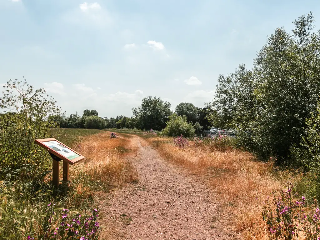 A path leading through a meadow. There is an information board on the left.