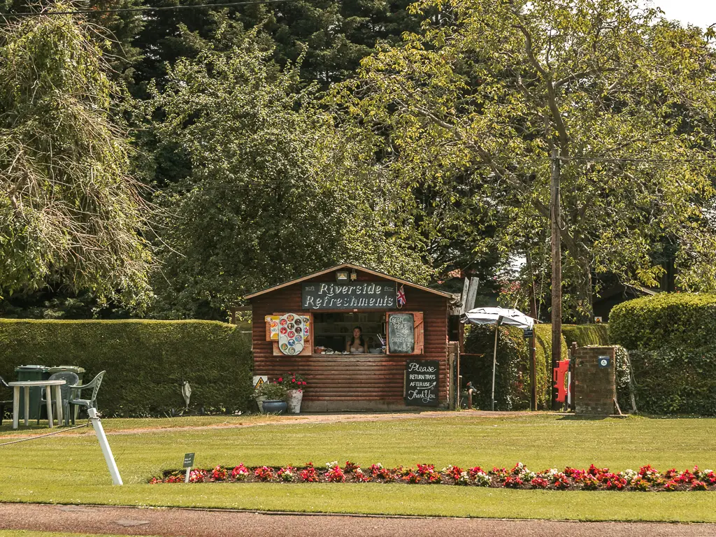 A wooden refreshment hut on the other side of a neatly cut green, with a row of red flowers. There is a hedge on either side of the hit and big trees behind.