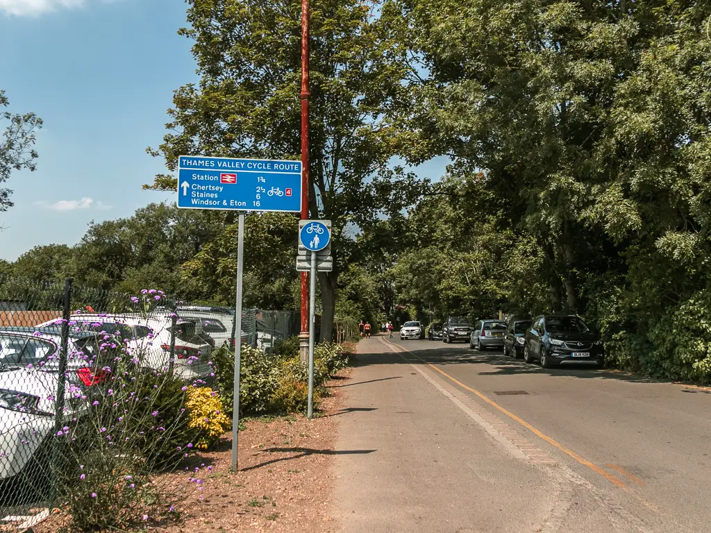 A road leading ahead with pavement on the left and cars parked behind a metal fence. There is a line of cars parked along the right side of the road as well. There is a big blue sign on the left side of the pavement.