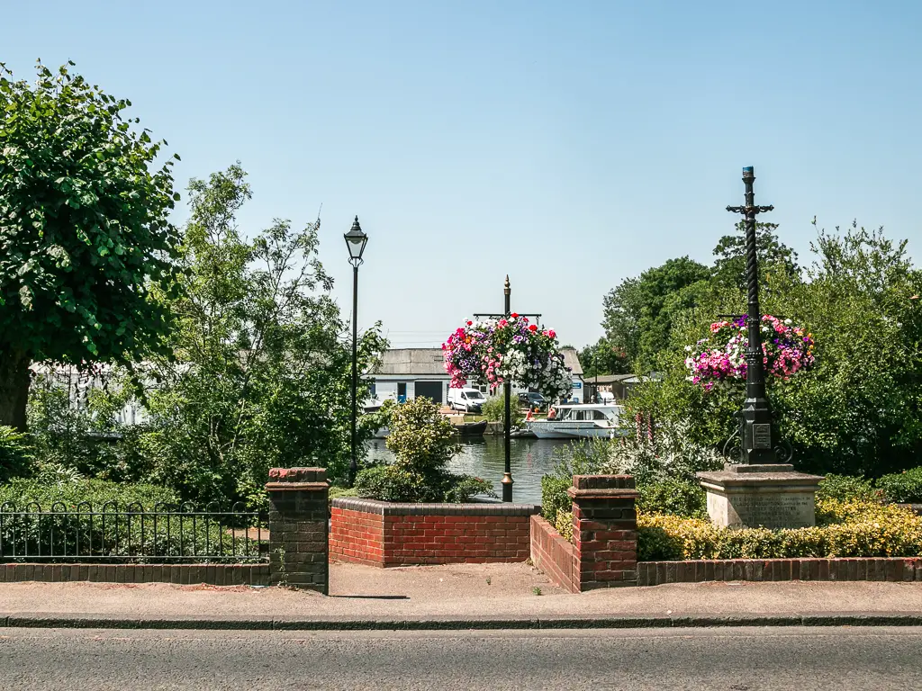 Looking across the road to the path through the short brick wall towards the river,. There are hanging flower baskets and a few bushes and trees on the other side of the wall.