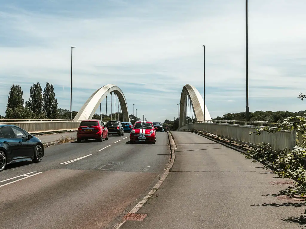 A road and pavement leading over a bridge with lots of cars driving.