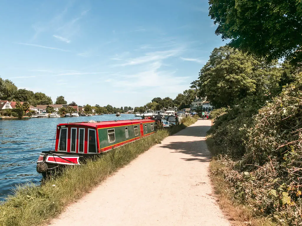 A long pale path leading ahead, on the walk from Shepperton to Hampton Court. The river is to the left with barges moored to the side. There are green leafy bushes on the right.