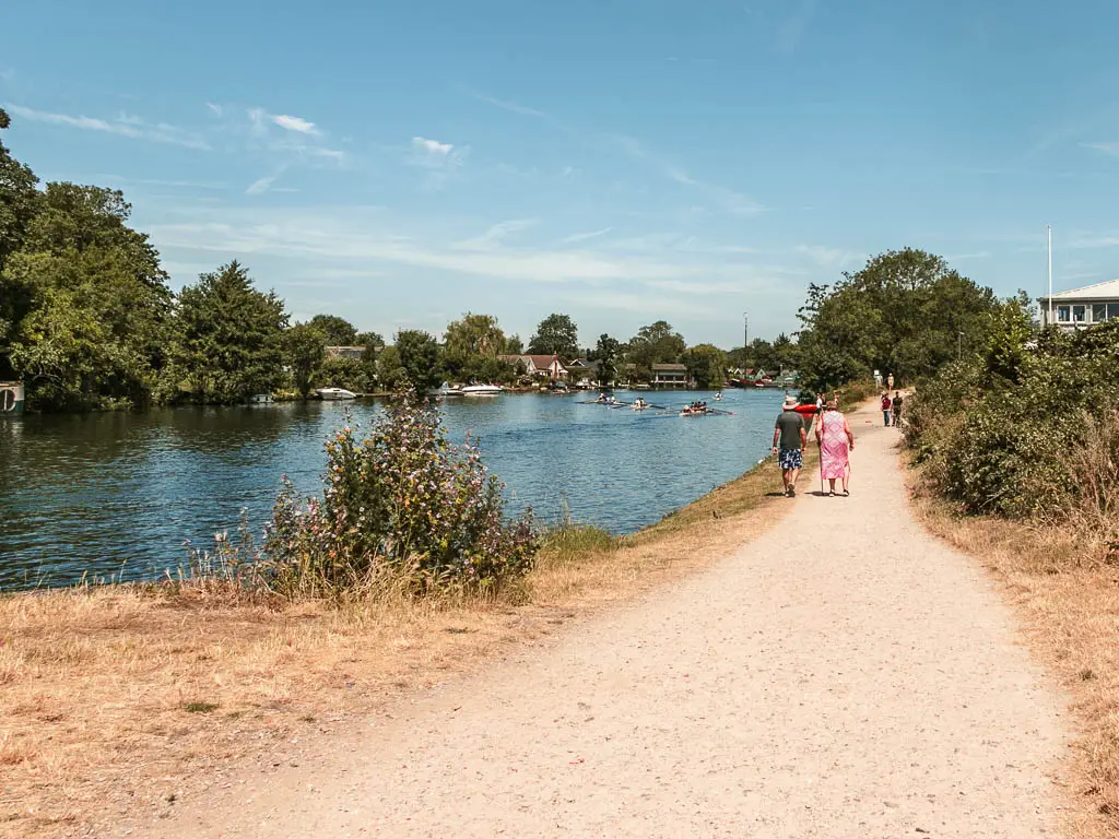 A long straight gravely wide path leading ahead, with the blue river to the left, when walking between Shepperton and Hampton Court. There are people walking along the path ahead.