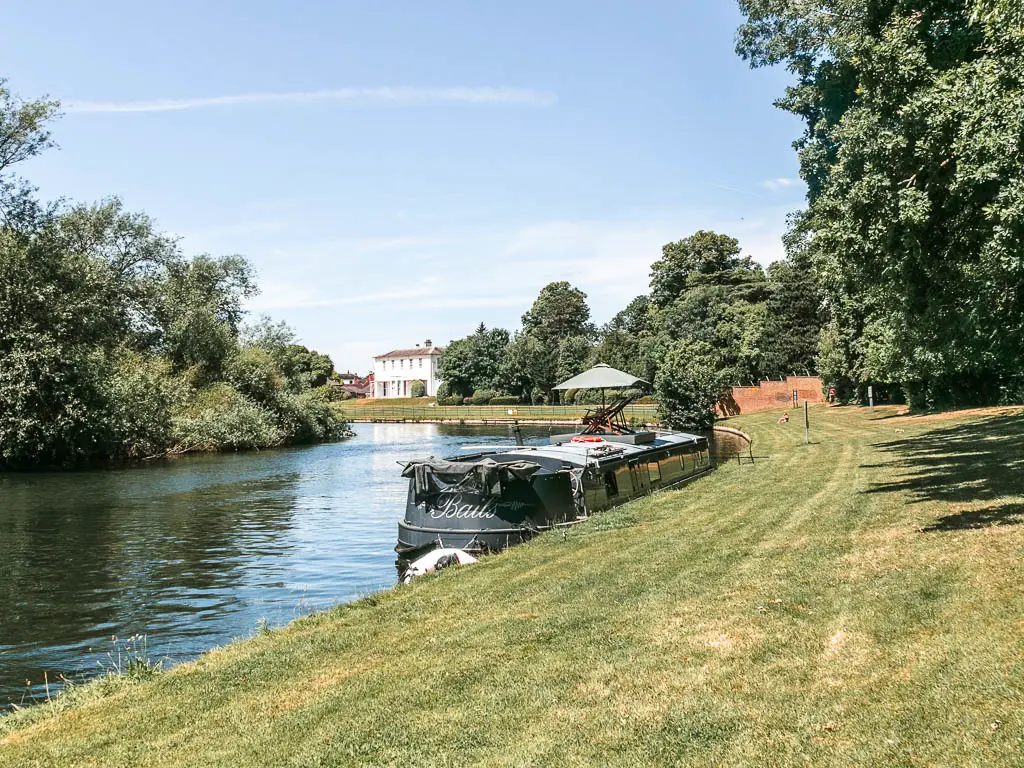 A neatly cut wide area of grass, with the river to the left and barge moored to the side at the start of the walk from Shepperton to Hampton Court. There is a large white walled house visible in the distance. 