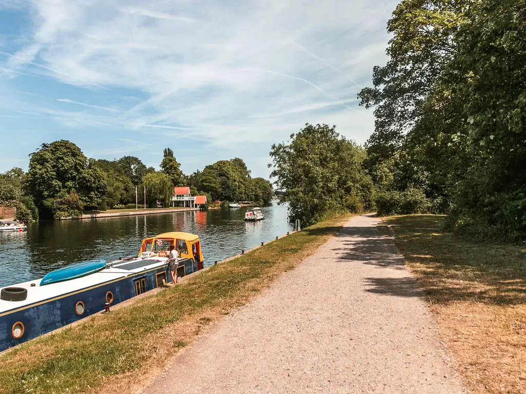 A wide gravel type path leading ahead, lined with strips of grass. The river is to the left and a blue barge is moored to the side.