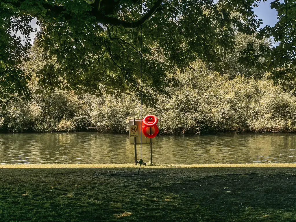 Looking over the green under tree shade, to the river. There is a red flotation ring on a stand by the river. There other side of the river is lined with trees.