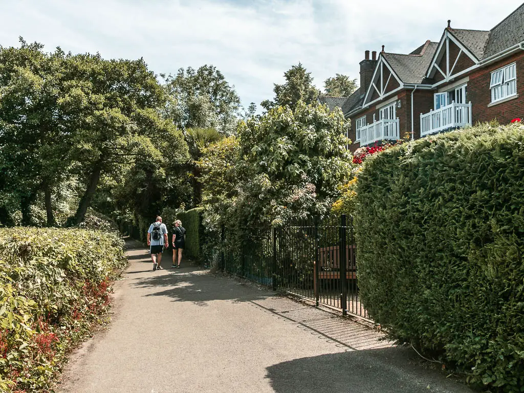 A with path lined with hedges on both sides, and the tops of houses visible to the right. There are two people walking on the path ahead.