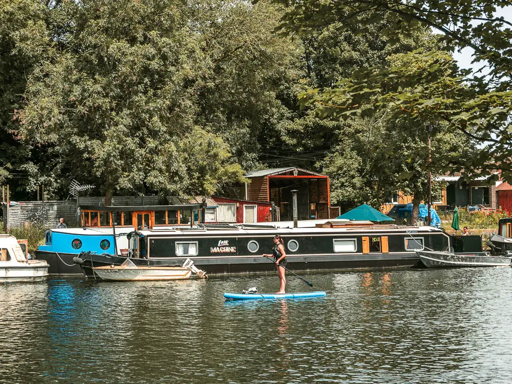 A woman on a stand up paddle board in the river. There is a black barge moored to the side behind he.