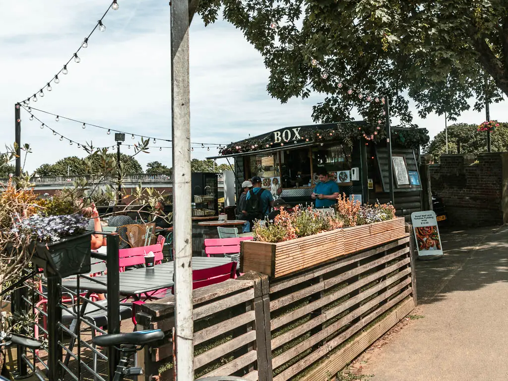 Looking over a wooden fence to a pop up black coffee hut in Hampton court. There are a few people standing outside the coffee hut and tables and chairs. 