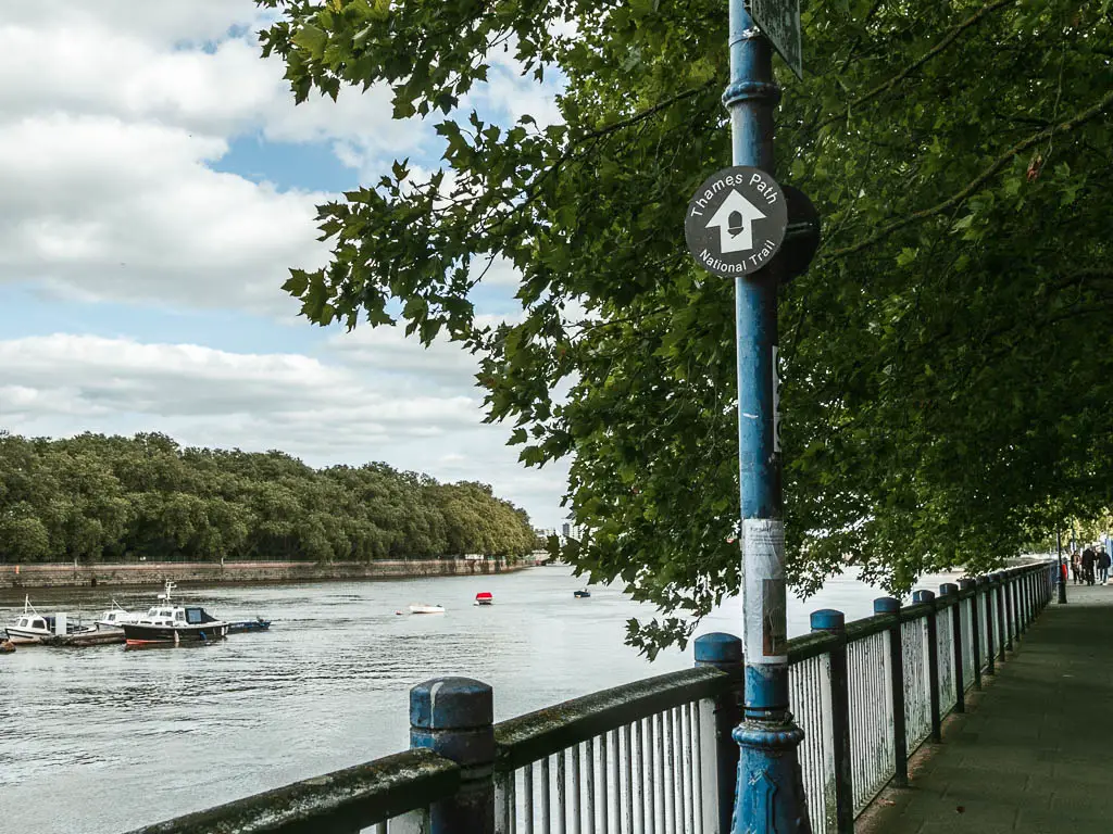 A black circular Thames Path sign on a blue lamppost pointing ahead. There is a metal raining to the  left with the river on the other side. There are a few boats on the river, and masses of green leafy trees on the other side.