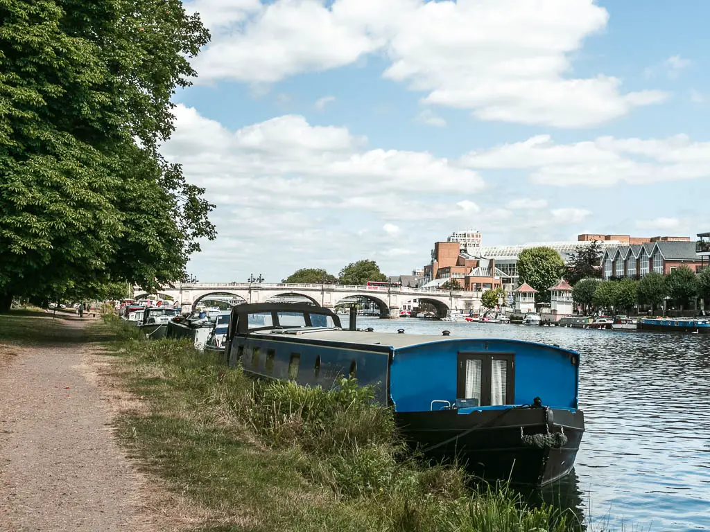 A dirt walking path on the left with strip of grass then rip er to the right. There is a blue barge moored to the side, and a bridge ahead.