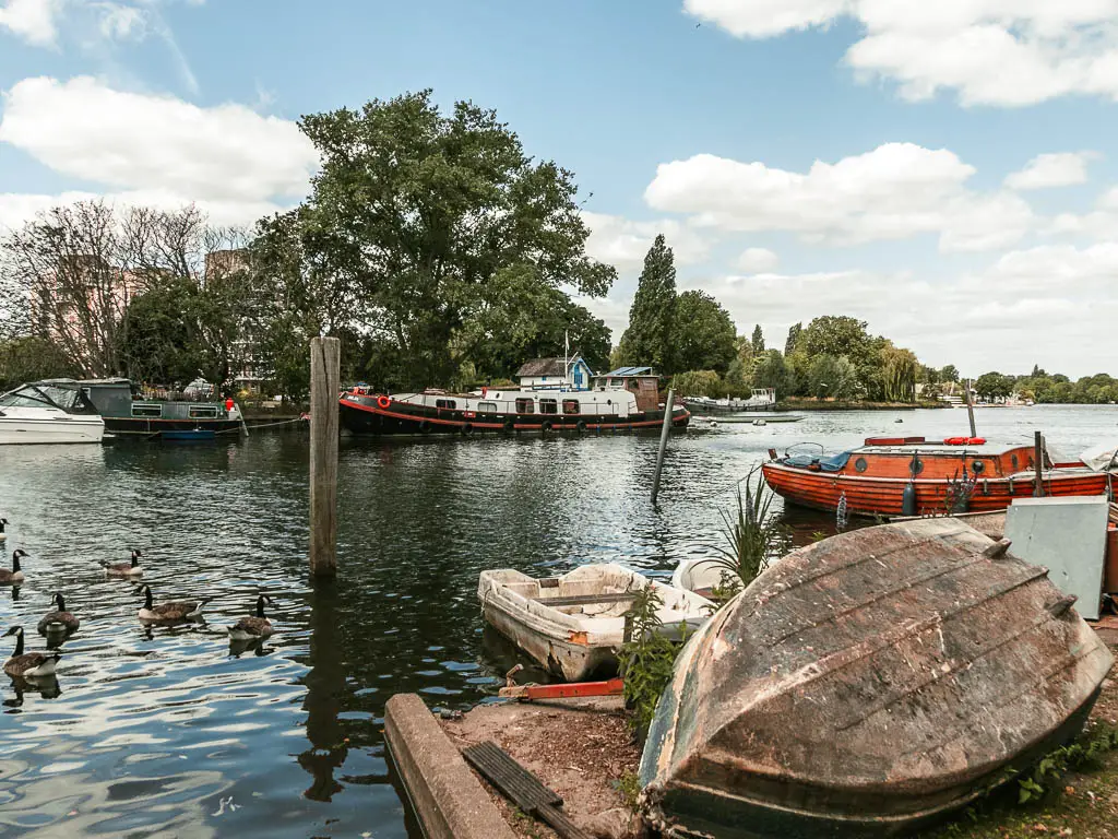 Looking over an upturned small boat next to the river, when walking between Hampton Court and Putney.  There are some boats and trees on the other side. 